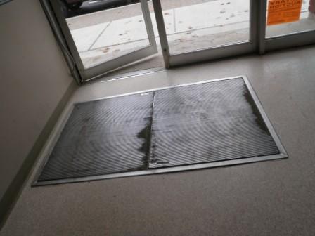 .Here we have a photo from the Orillia home inspector.biz evidence files- flooring section. This dirt catching screen let into the floor is commonly found in commercial inspections. It and others like it are used to trap moisture, dirt, and debris, from foot traffic before it tracks through the building. Most of these have removable screens on top so they can be cleaned out. Most have drains so they can be flushed out as well. The better units have a stainless steel lined holding box under the screens. I have also seen painted steel, rubbe,r and just recently a plastic pan liner. Around Orillia the best units I have seen on commercial inspections are built with a wide flange that sits flush on the sub floor and a second flange that allows the screen to sit flush to the finished floor.  This type allows a good seal from the finished floor to the screen flange.  But what if the pan is not made this way?  See the next photo. 